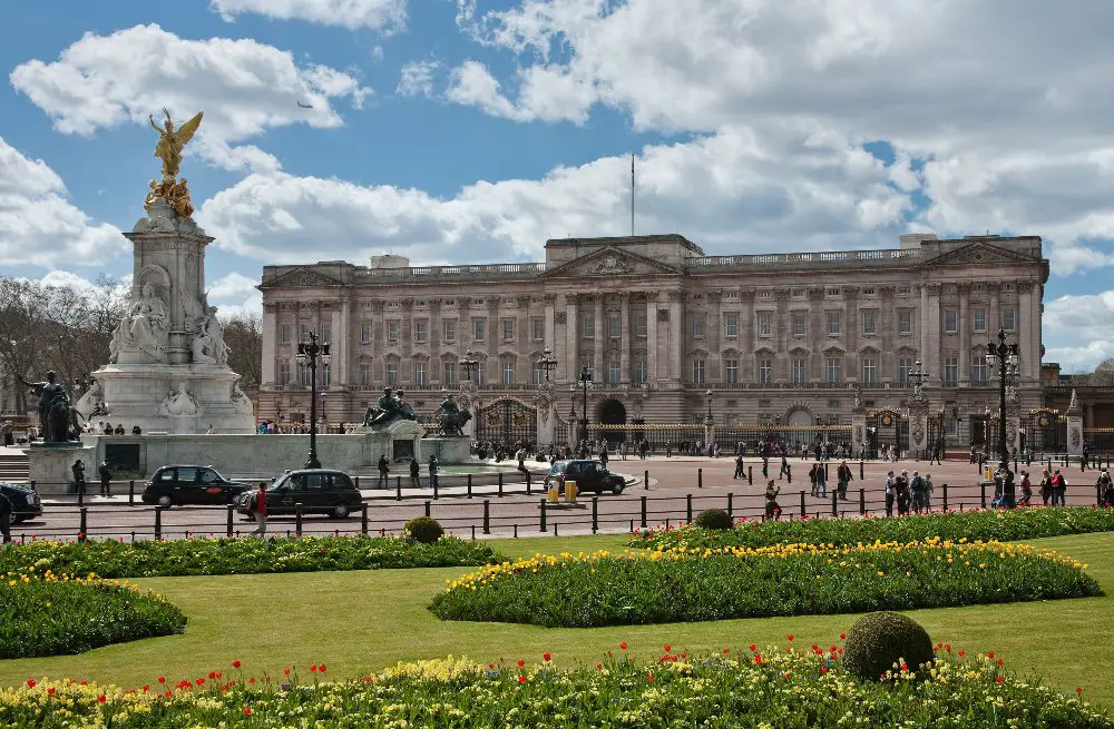 view of buckingham palace in the distance with lawns in front, black cabs and people walking around, a statue in front of the palace, and a cloudy sky