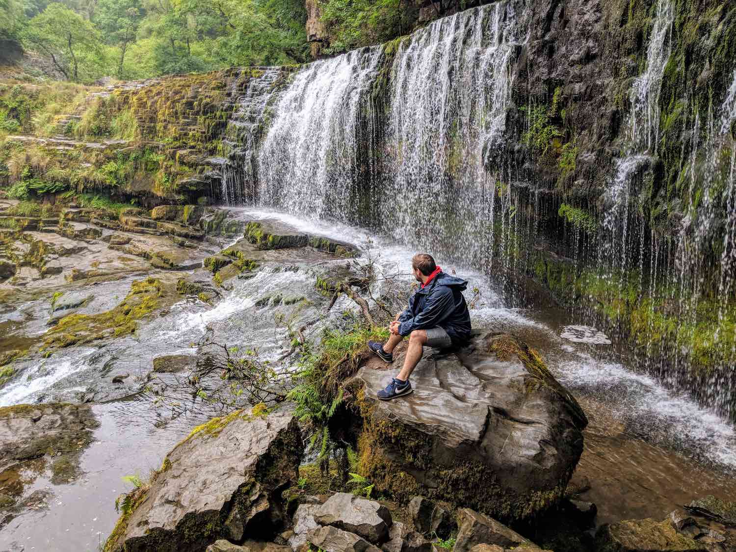 man sitting on a rock in front of a wide waterfall at the brecon beacons, wales