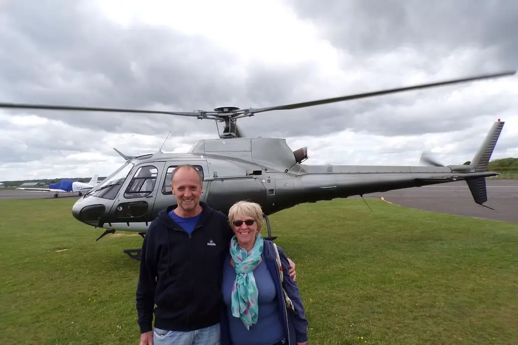 a happy couple standing and smiling in front of a parked helicopter at a heliport