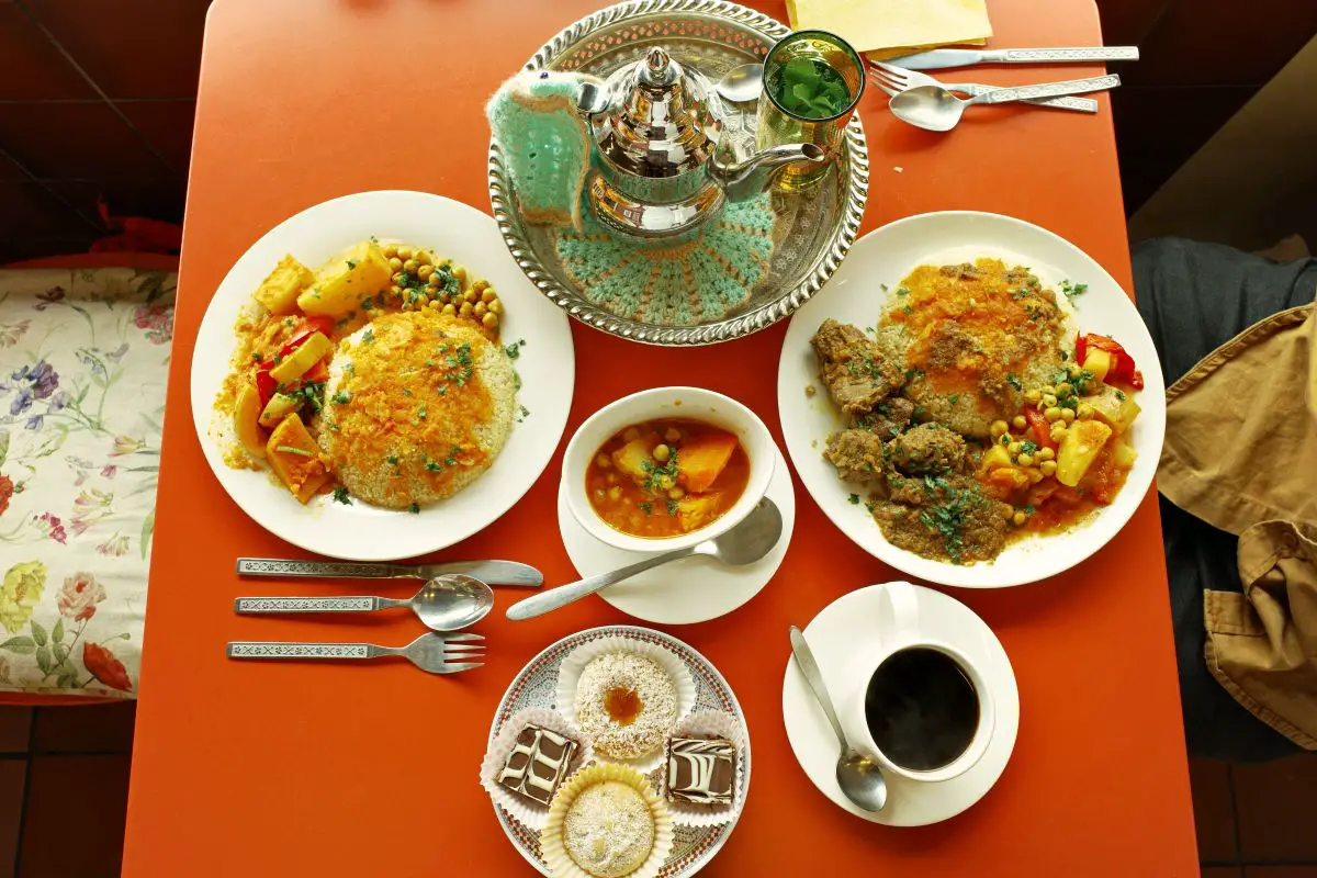 birds eye view of the main dishes on a red table consisting of vegetable tagine, lamb and couscous, vegetable couscous, moroccan mint tea, moroccan coffee, and traditional sweets