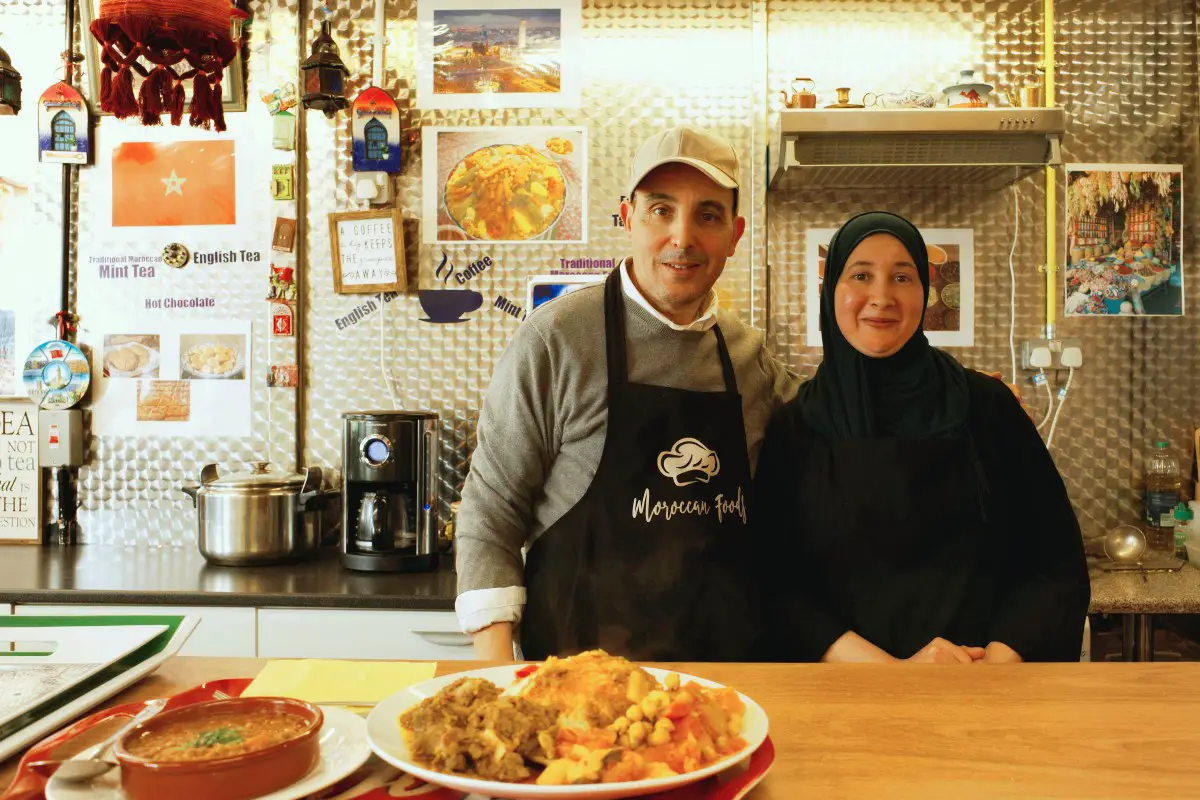 a moroccan couple in their 30s-40s smiling for the camera at the front of their moroccan foods stall in stoke, england