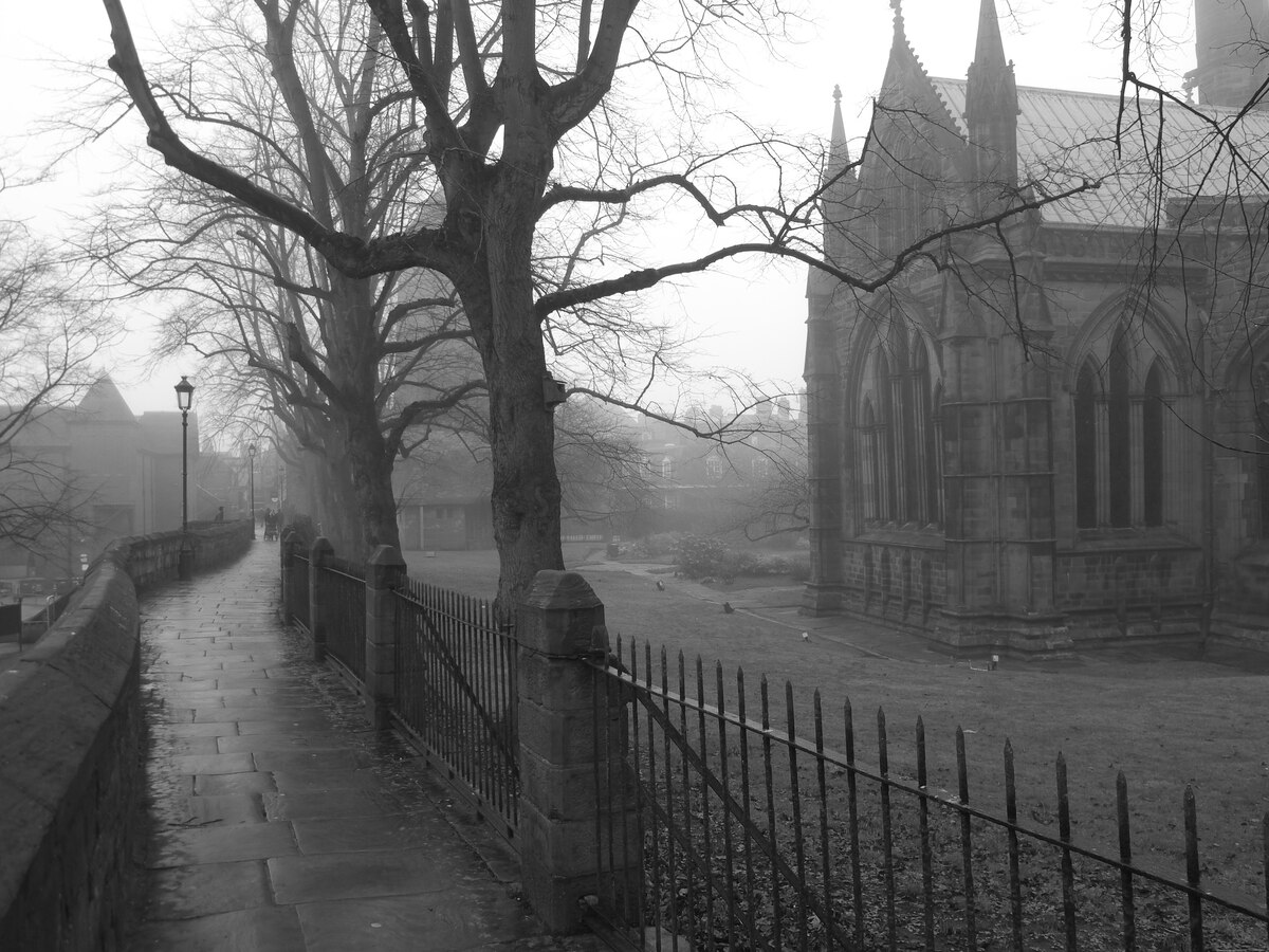 A black and white image of a misty path alongside a Gothic church. Bare trees line the path, which is bordered by an iron fence. The spires and arched windows of the church are visible through the fog, creating a moody atmosphere.