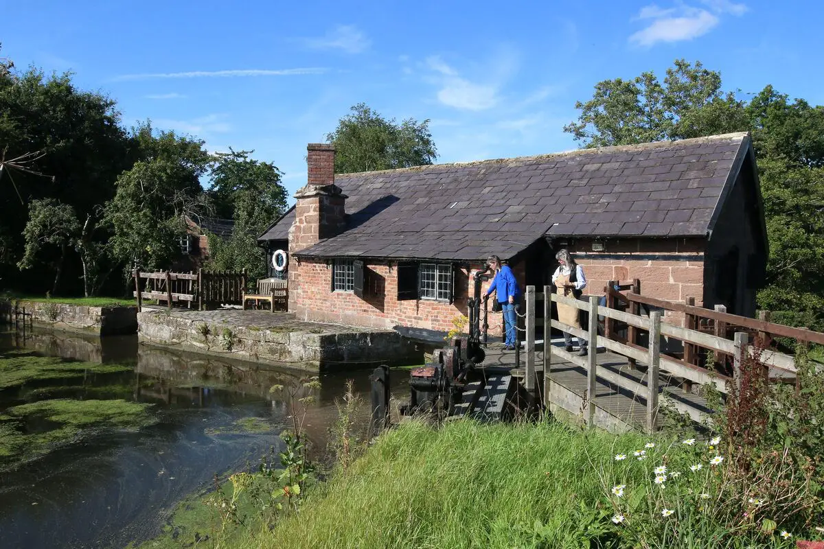 A rustic brick building beside a canal or small river. The building has a slate roof and appears to be an old watermill. There's a wooden footbridge leading to the building, and two people can be seen on it. The surrounding area is lush with trees and grass, and there are some wildflowers in the foreground. The sky is blue with a few clouds.