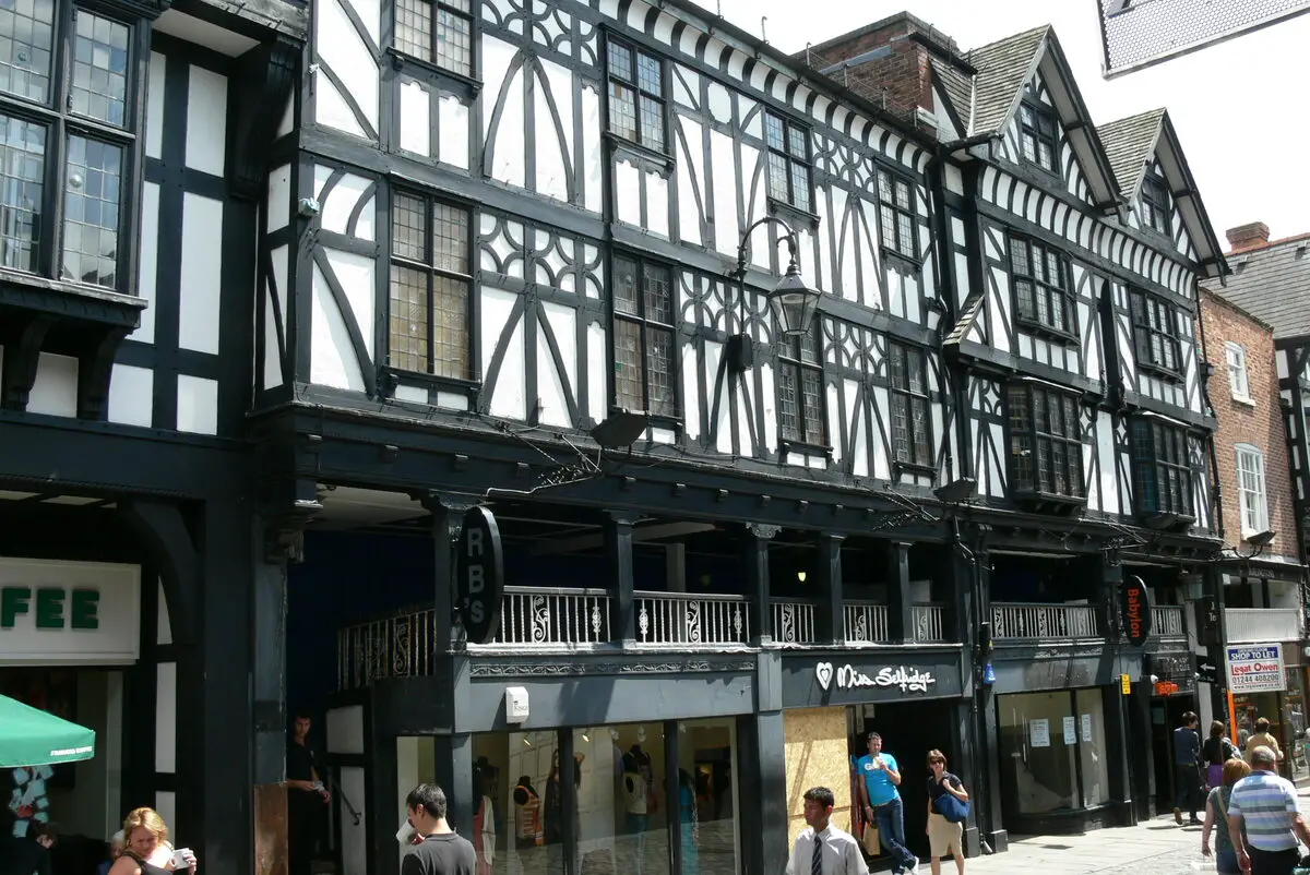 A Tudor-style building with black and white timber framing in Chester, England. The building has overhanging upper floors supported by decorative brackets. The ground floor contains shops with modern storefronts. Pedestrians can be seen walking along the street in front of the building.