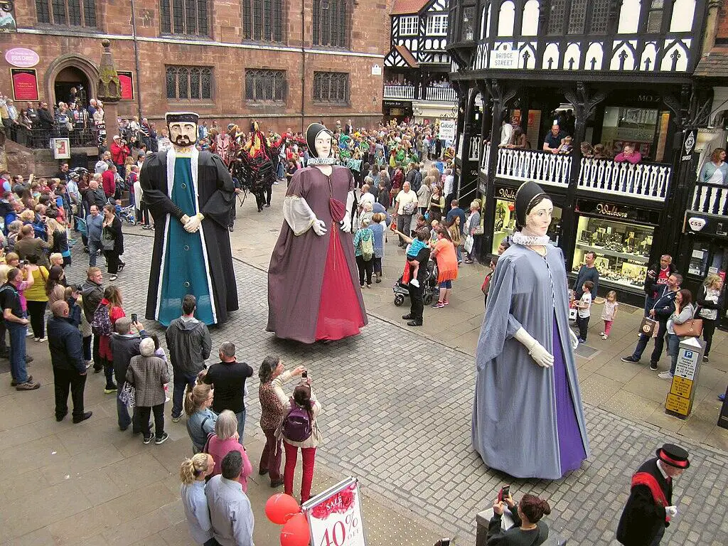 A crowded city square with historic Tudor-style buildings. Three giant figures in medieval costume tower over the crowd. People gather around, taking pictures and watching the spectacle.