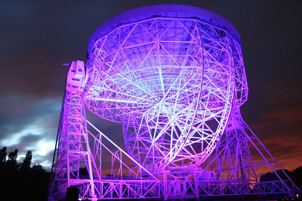 A large radio telescope illuminated in vibrant purple and pink lights against a dark night sky. The telescope's dish and supporting structure create an intricate lattice of metal beams.