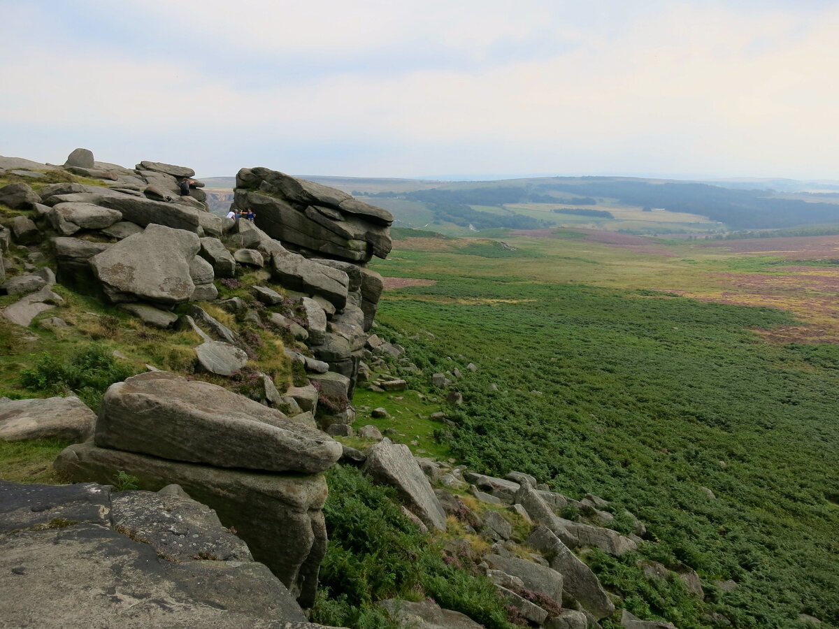 A rocky outcrop overlooking a vast moorland landscape. Large, weathered boulders are in the foreground, with green and purple heather-covered hills stretching to the horizon. A hazy sky is visible in the distance.