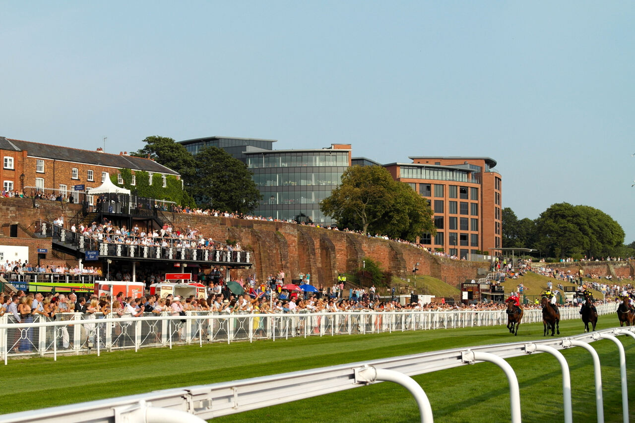 A horse racing track with horses running on the grass. Spectators watch from behind a railing and from elevated stands. Modern office buildings are visible in the background behind old brick walls.